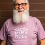 An older man with a white beard wearing a classic purple t-shirt.