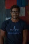 A man with curly hair and glasses wears a dark "Vote Unmatrix" t-shirt. He stands against a colorful, patchwork background that adds a touch of vibrancy to his trendy, humorous apparel.